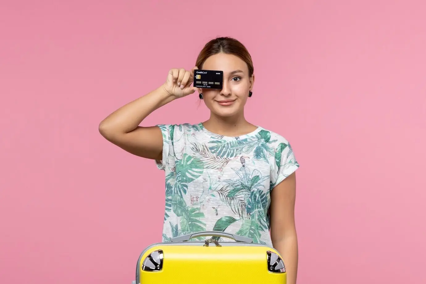 Frontal view of a young woman holding a black bank card in front of the pink wall.