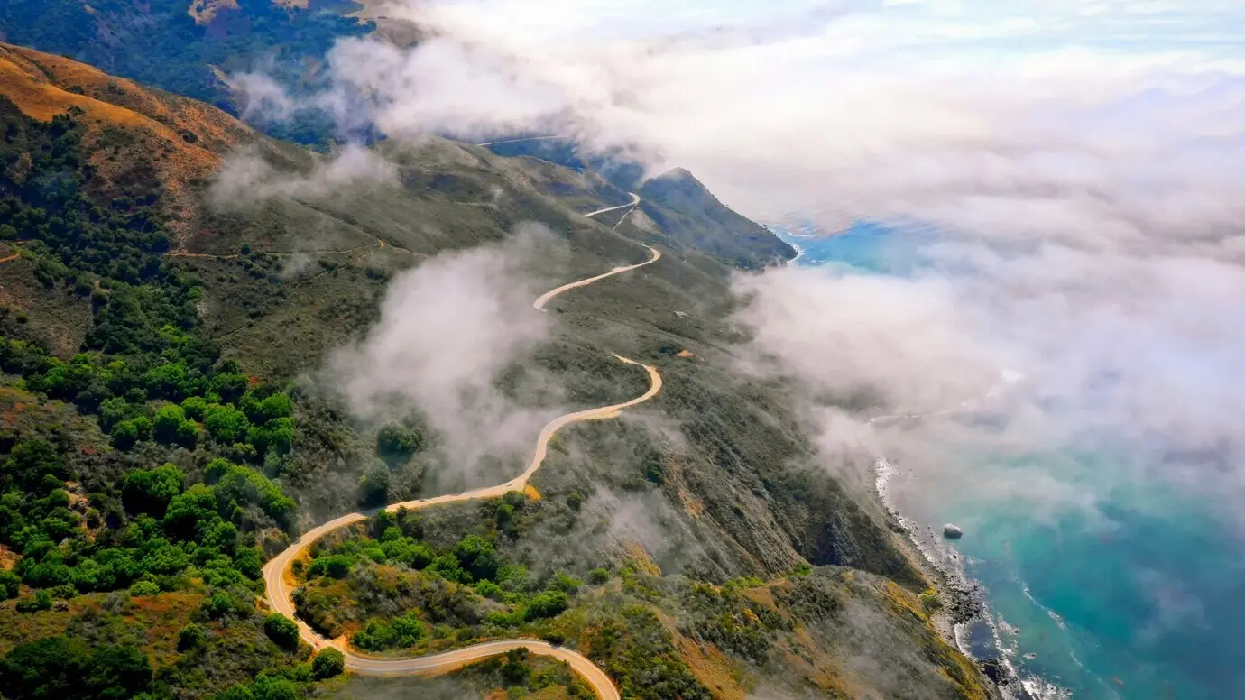 An aerial shot of beautiful green hills with a curving road along the edge and an amazing sea.