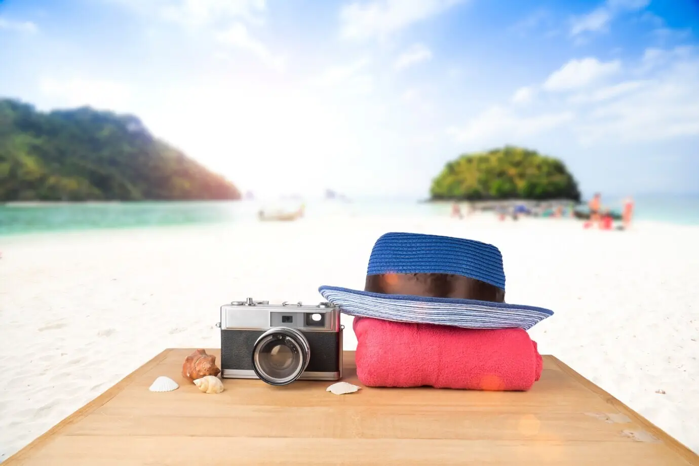 A red-pink tower, a blue hat, an old vintage camera, and shells on a wooden floor with a sunny blue sky and ocean in the background.