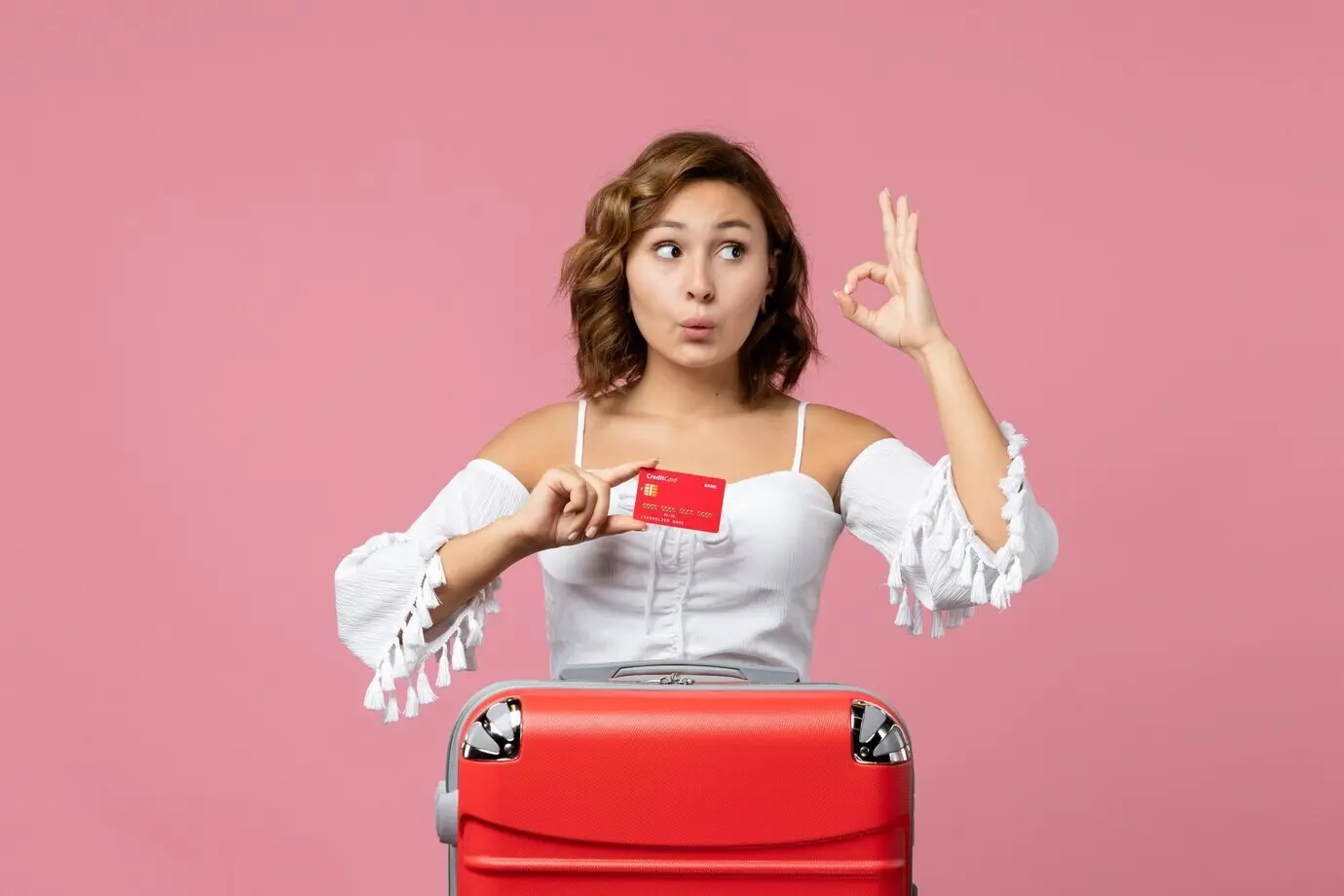 A front-facing view of a young female model on a pink floor, with a vacation bag and holding a bank card; trip, vacation, color, voyage, sea.