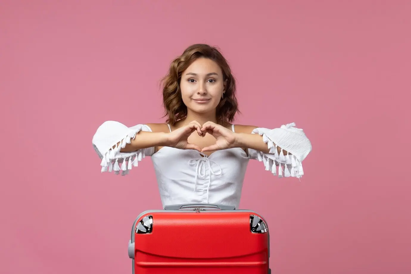 Front-facing view of a smiling young woman preparing for a trip with a red bag in front of a pink wall.